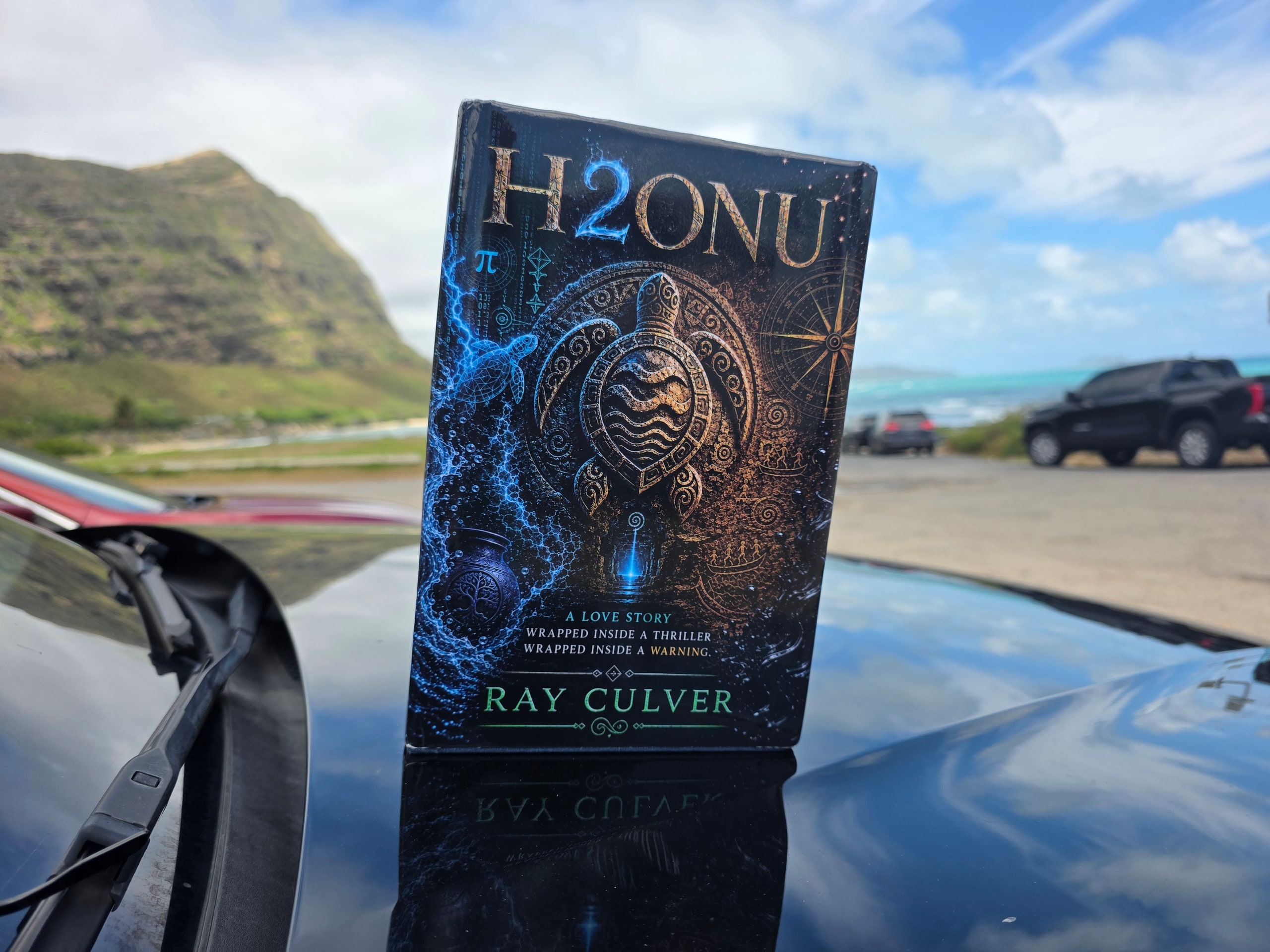 H2ONU First Edition hardcover on a car hood, turquoise water and mountain behind